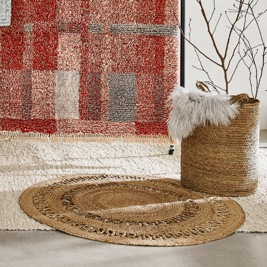Ovaler Jute-Teppich in Naturfarbe auf hellem Teppich mit rotem Muster im Hintergrund